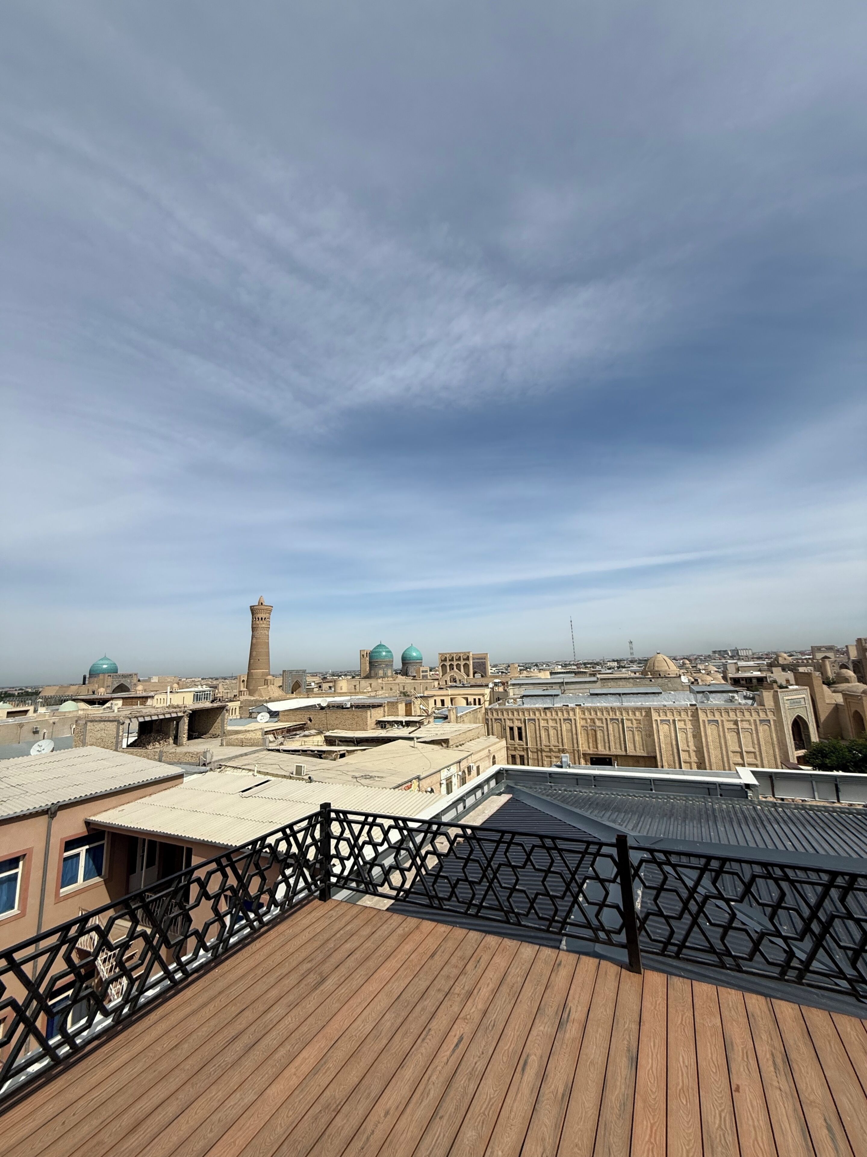 Une terrasse sur le toit, avec un sol en bois et une rambarde en métal, offrant une vue sur le paysage urbain, avec ses bâtiments historiques et sa tour.