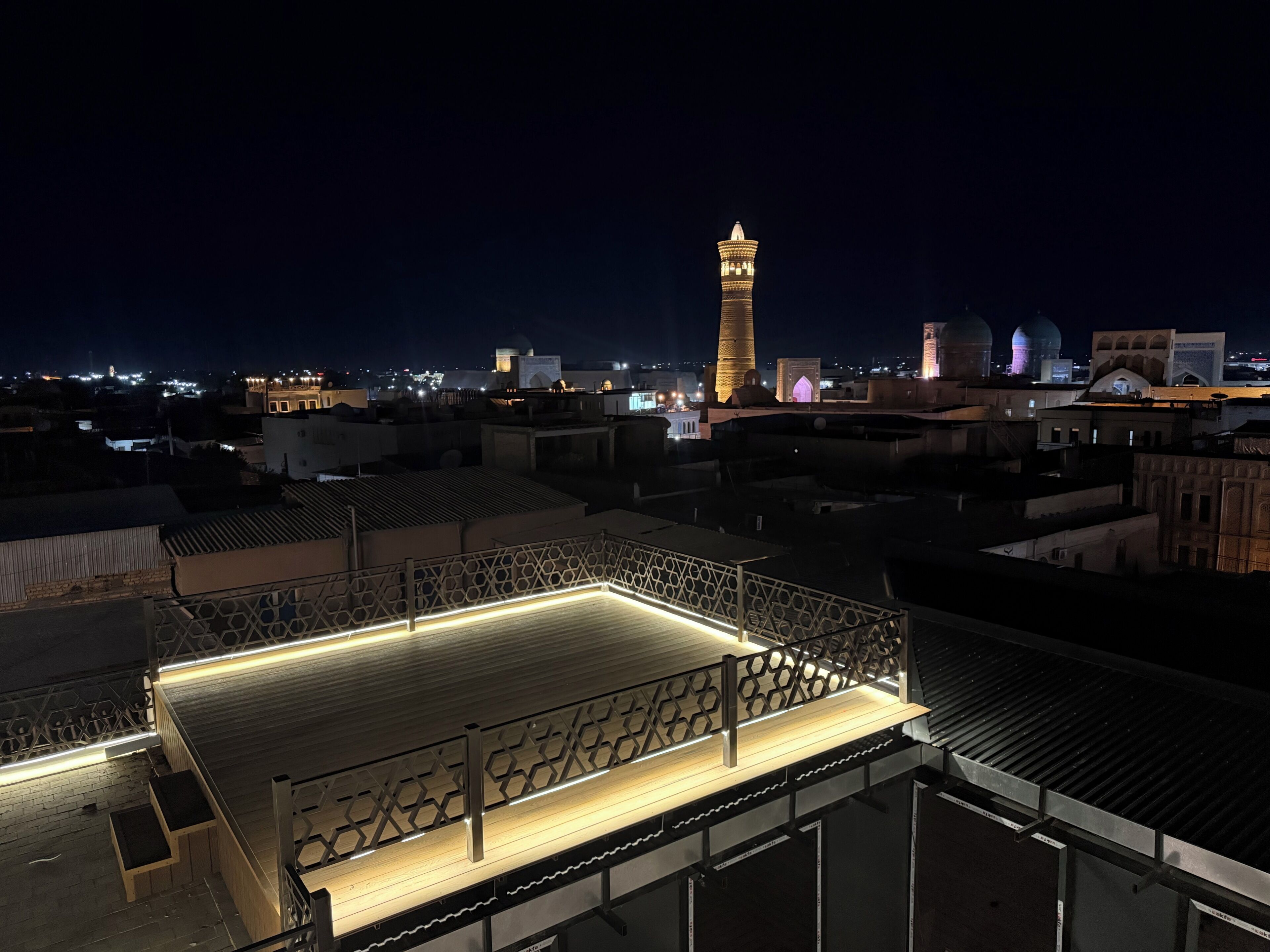 Una terraza en la azotea con barandilla, vista panorámica de la ciudad con edificios iluminados al fondo y una torre destacada con cúpula.