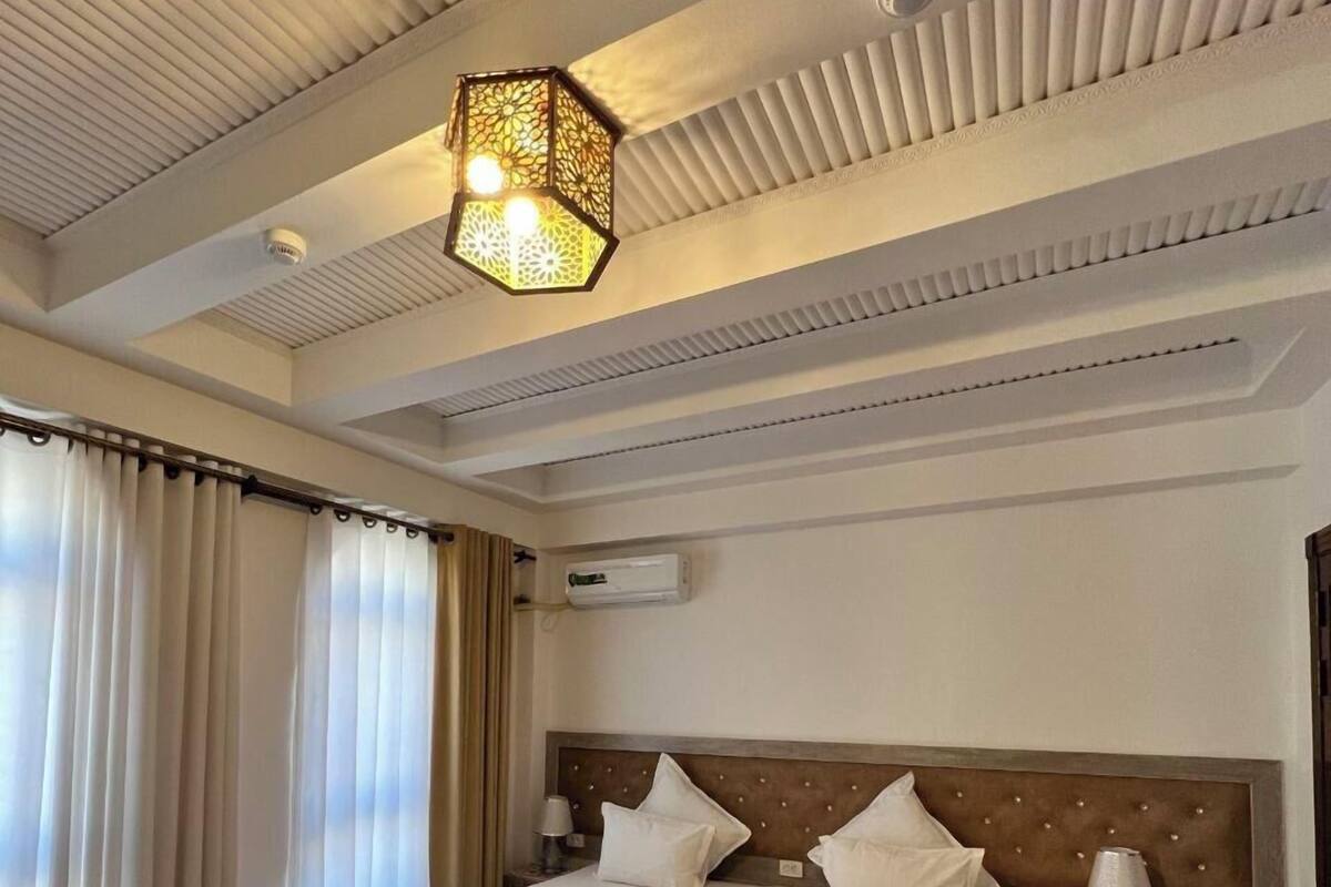 Ein Hotelzimmer mit einem großen Bett, einer gemusterten Tagesdecke, einem roten Teppich, einem Deckenventilator und einer Klimaanlage.
