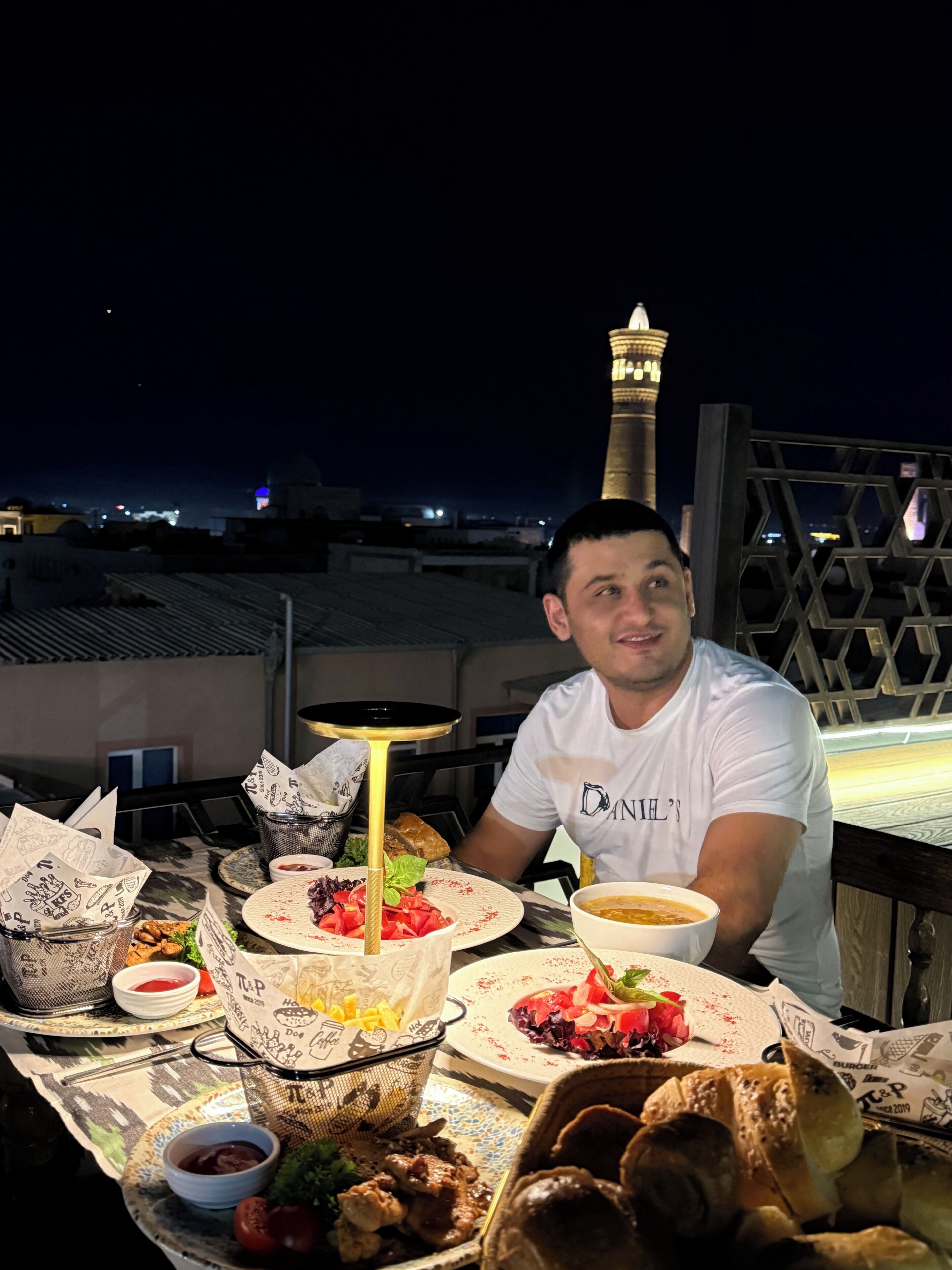 A man dining at an outdoor table with various dishes, a lamp, and a view of a tower in the background.