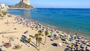 Une plage bordée de nombreux parasols et fréquentée par de nombreuses personnes, une formation rocheuse imposante et une mer d’un bleu limpide.
