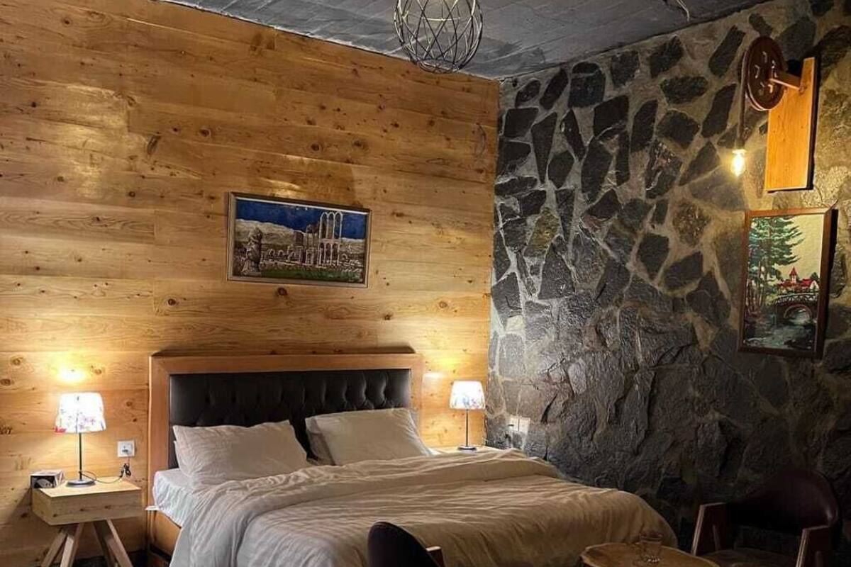 Une chambre confortable avec un mur en pierre, un plafond en bois et un lit doté d’une tête de lit. On y trouve deux tables de chevet avec des lampes, une chaise et une petite table avec une lampe.