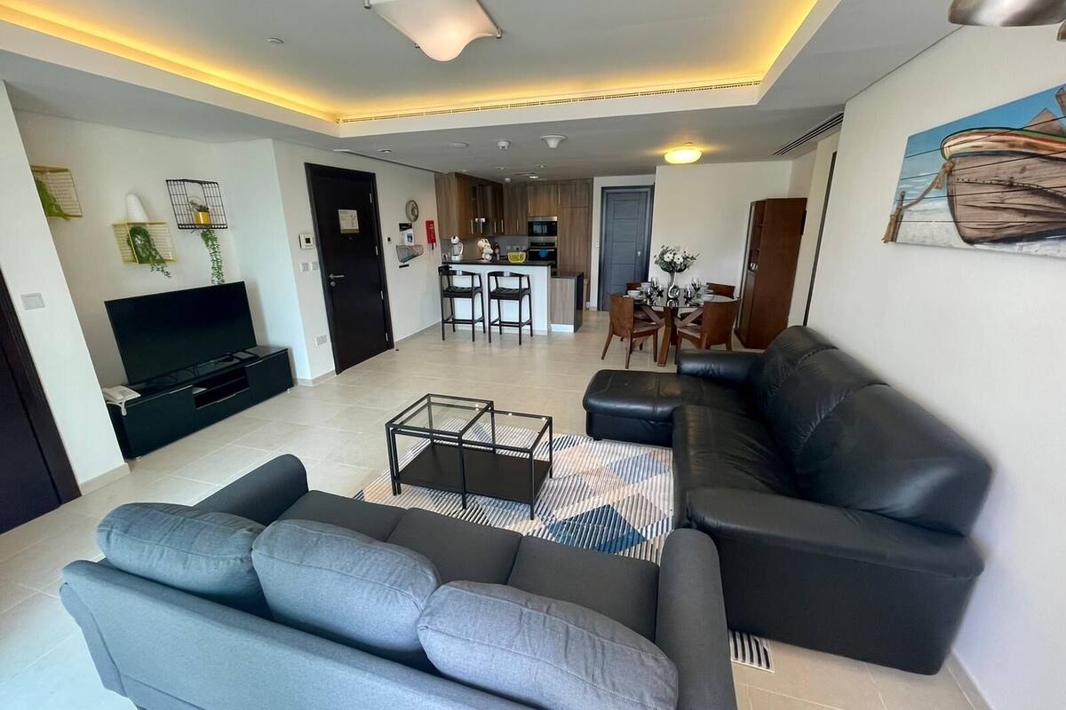 A modern living room with a sofa, a coffee table, and a TV. There is a kitchen in the background with a dining area.