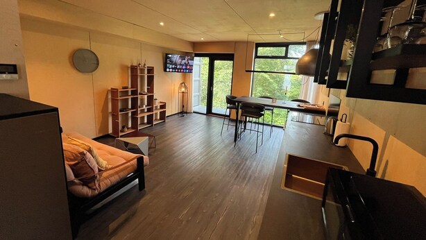Een moderne woonkamer met een bank, een eethoek met tafel en stoelen, een aan de muur bevestigde televisie en een trap naar een hoger niveau.