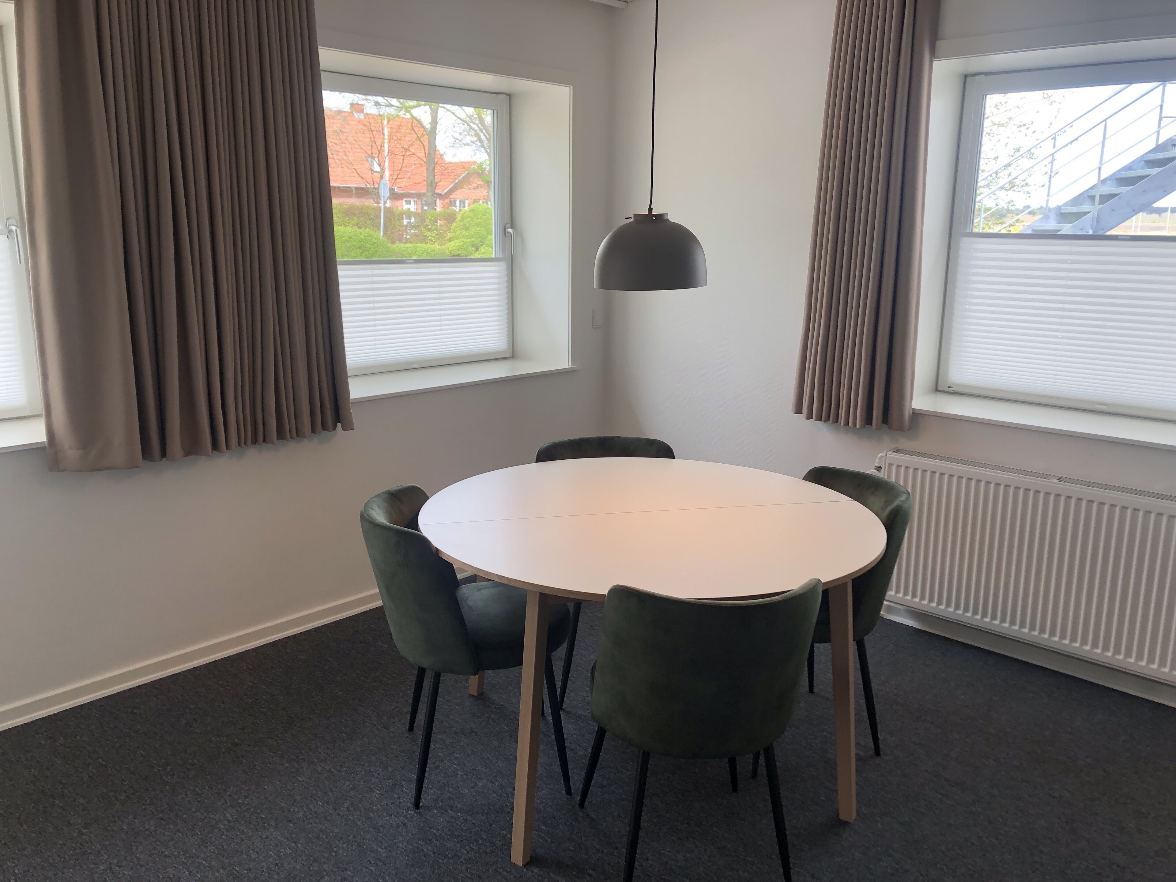 A modern dining area with a round table and four chairs, large windows with blinds, and a hanging pendant light.
