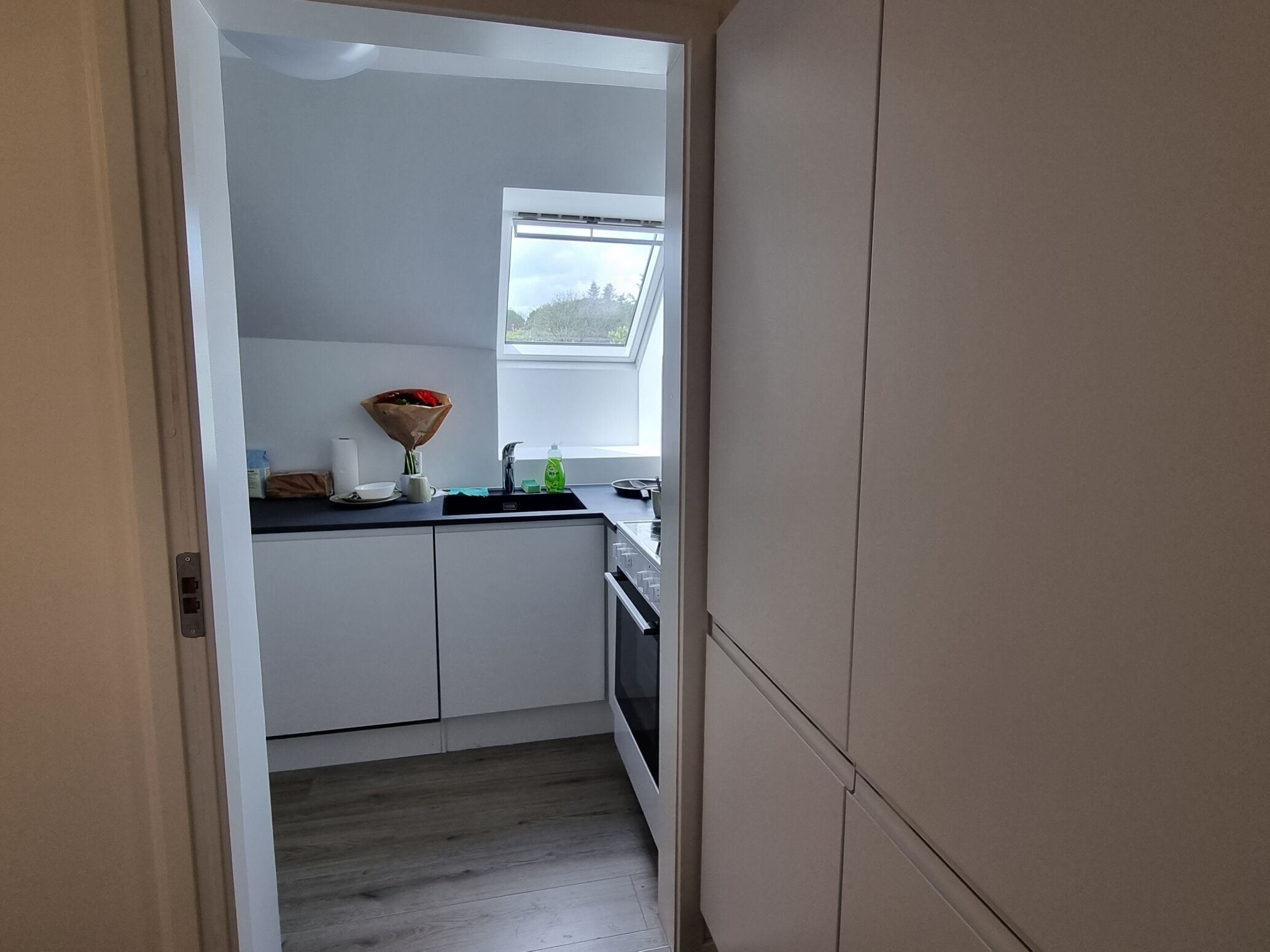 A narrow kitchen with white cabinets, a window, and a small countertop with a bouquet of flowers.