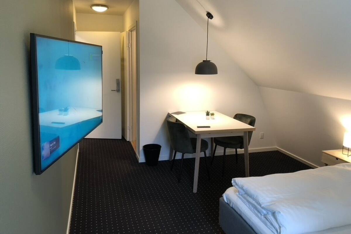 A hotel room with a bed, a desk with two chairs, a flat-screen TV mounted on the wall, a ceiling light, and a small table with a lamp.