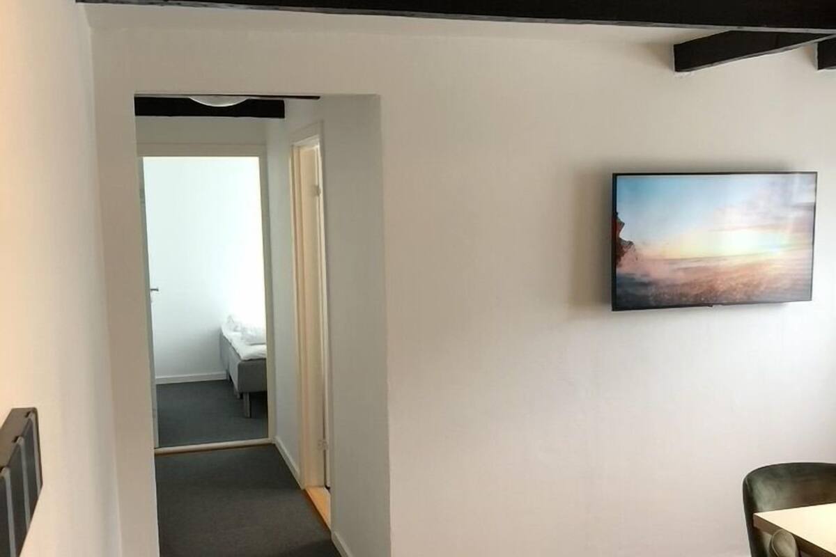 A hallway with a bed in the background, a painting on the wall, and a chair in the foreground.