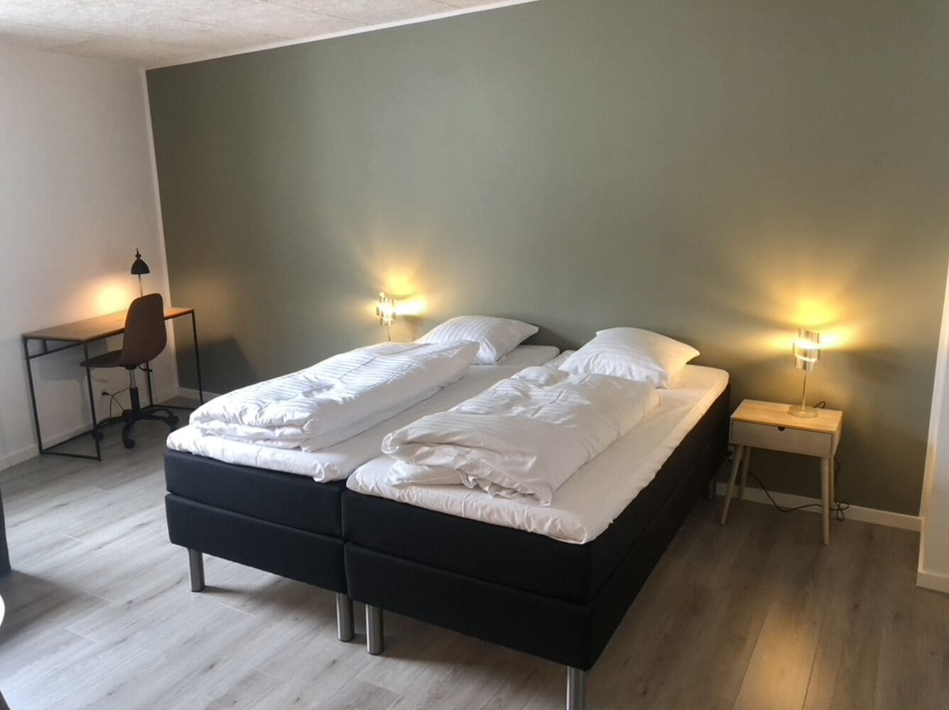 Una cama doble con ropa de cama blanca, dos mesitas de noche con lámparas, un escritorio con una silla y un suelo de madera de color claro.