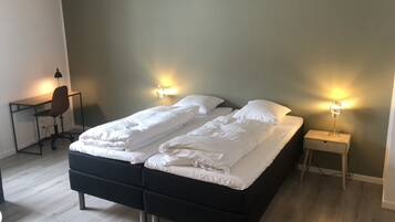 Una cama doble con ropa de cama blanca, dos mesitas de noche con lámparas, un escritorio con una silla y un suelo de madera de color claro.