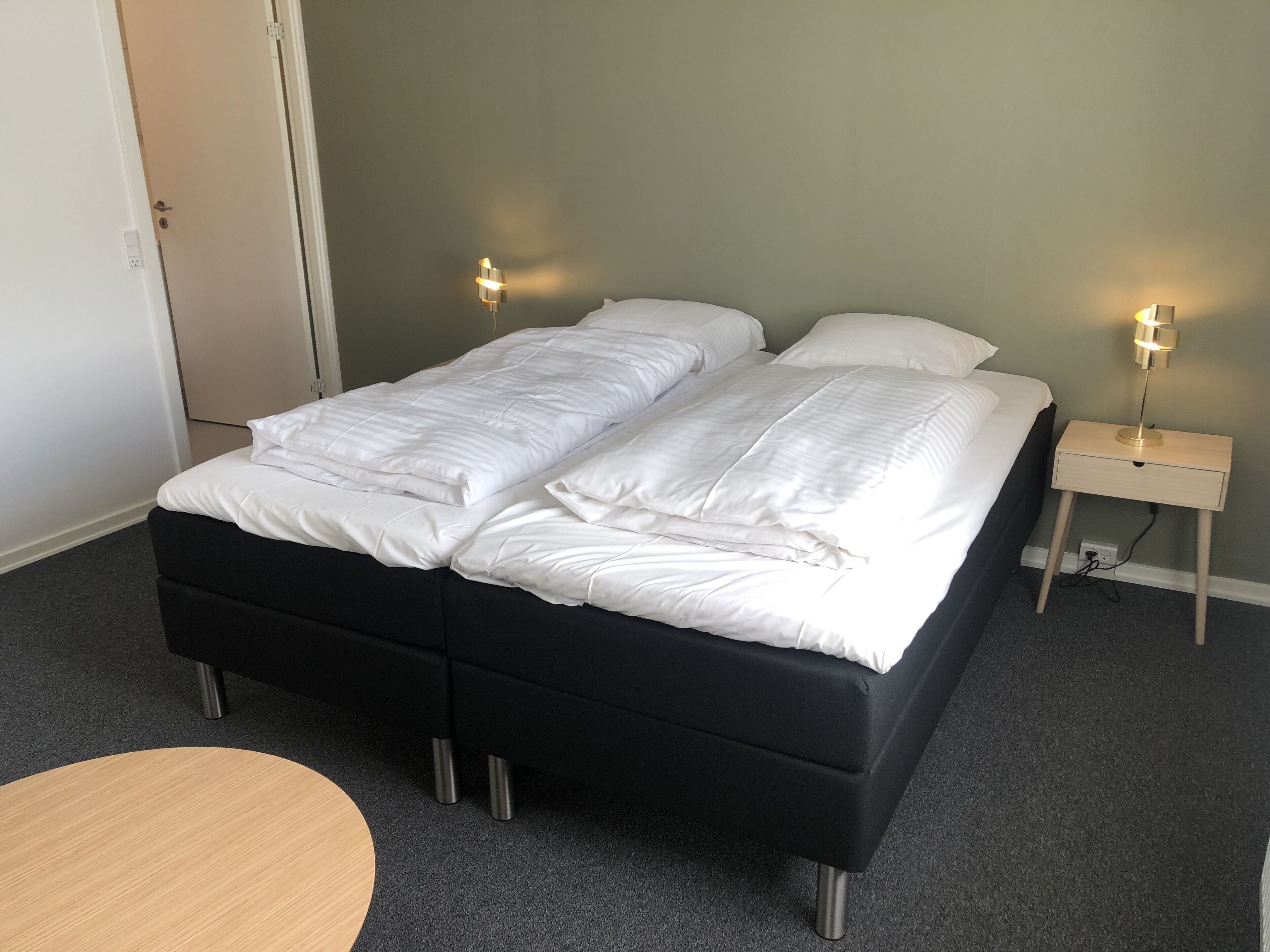 A double bed with white bedding, a small bedside table with a lamp, and a round wooden table.