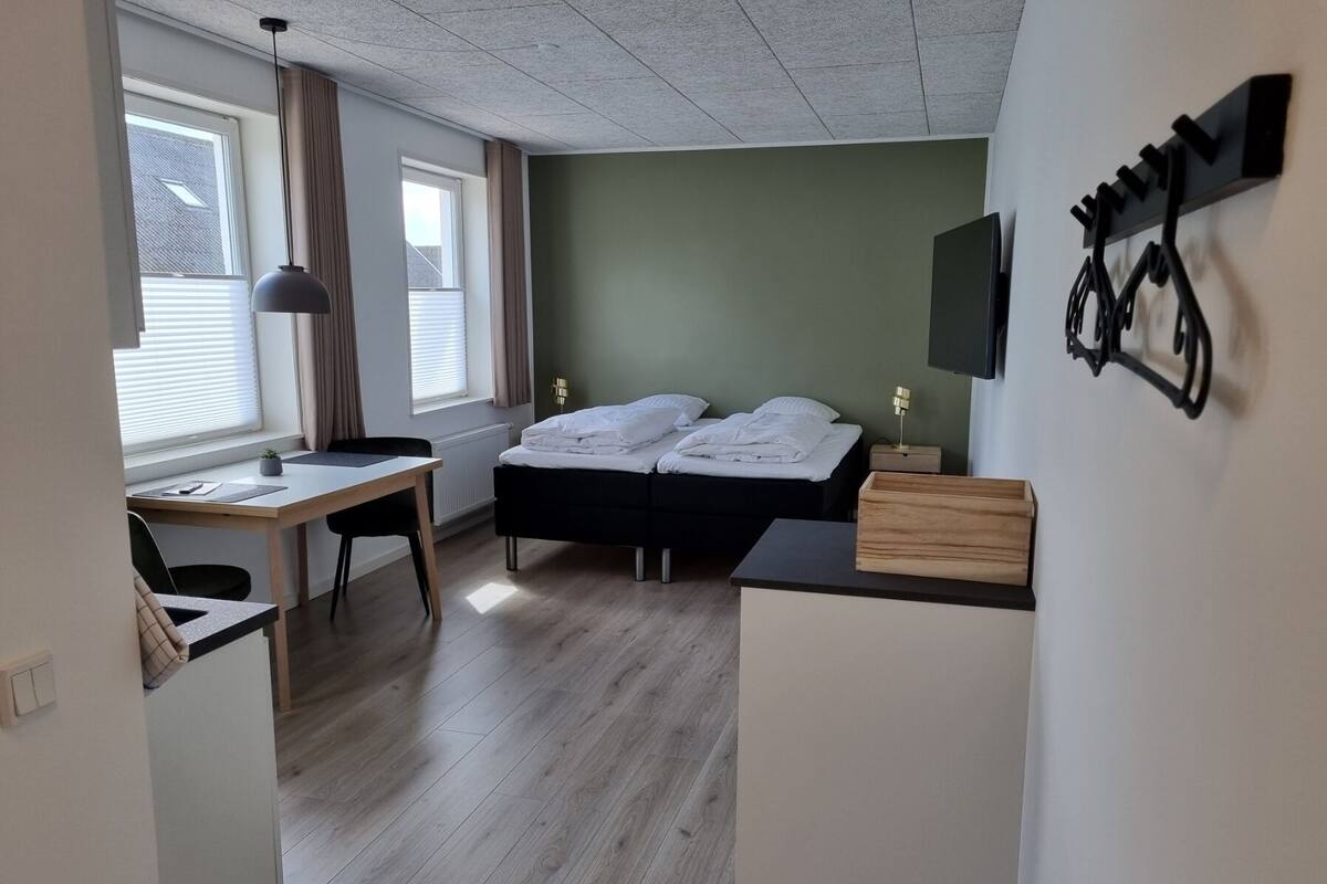 A compact room with a bed, a desk, and a chair. There is a window with blinds, a lamp, and a mounted TV.