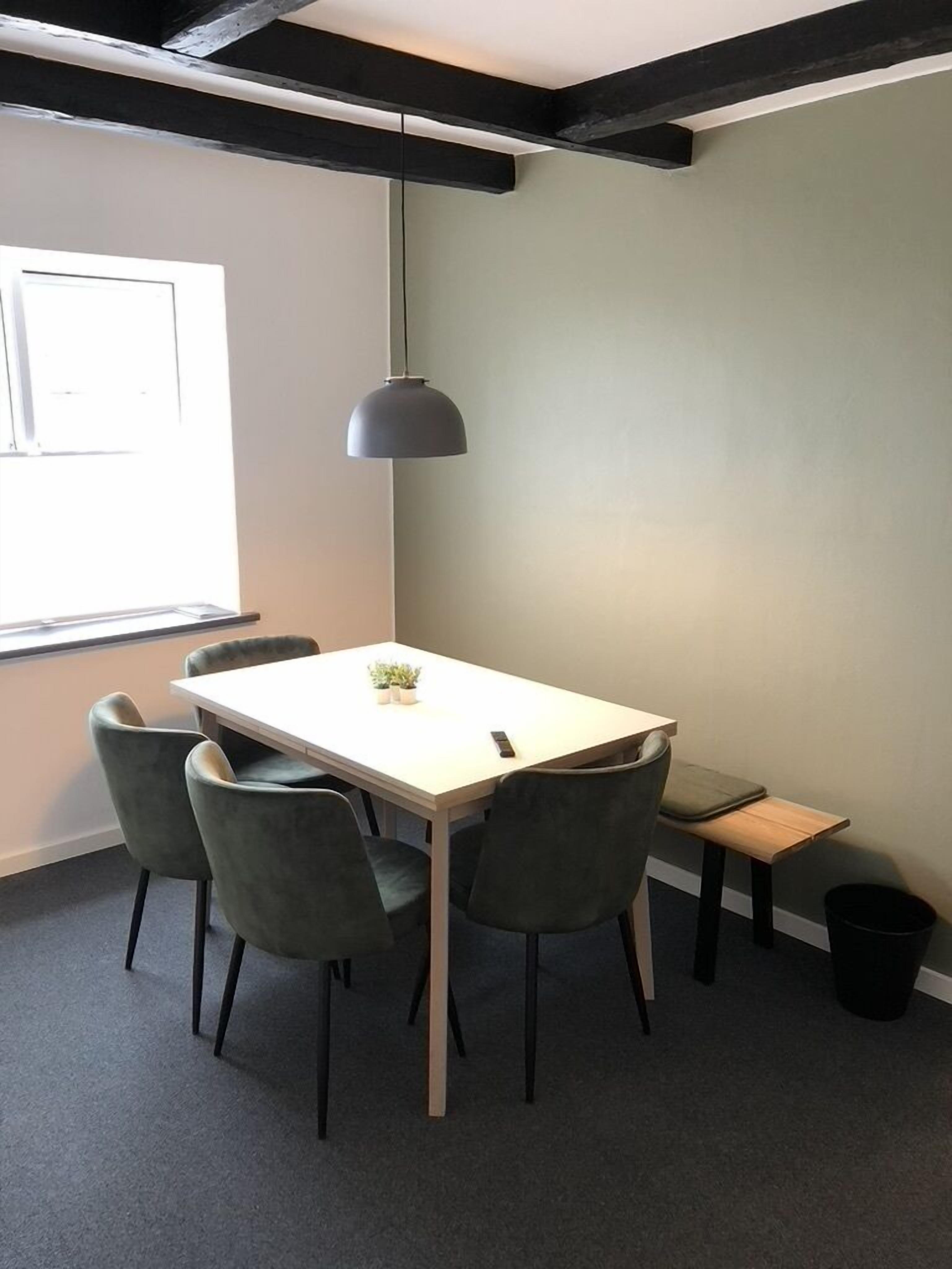 Una sala riunioni moderna con un tavolo bianco, quattro sedie grigie, una lampada a sospensione e una piccola panca di legno.