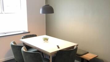 Una sala riunioni moderna con un tavolo bianco, quattro sedie grigie, una lampada a sospensione e una piccola panca di legno.