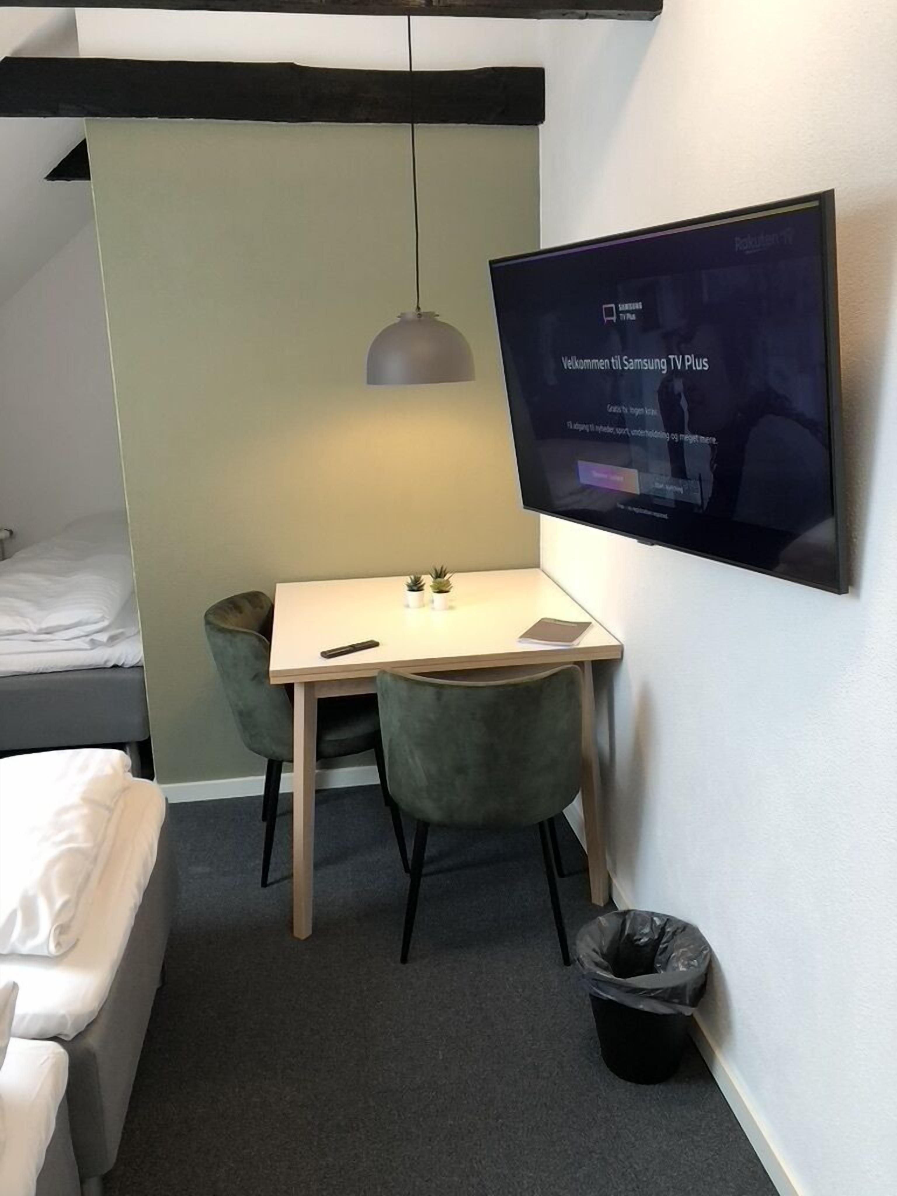 Habitació d'hotel petita amb un llit, un escriptori, dues cadires, un televisor fixat a la paret, una làmpada penjant i un safataire.
