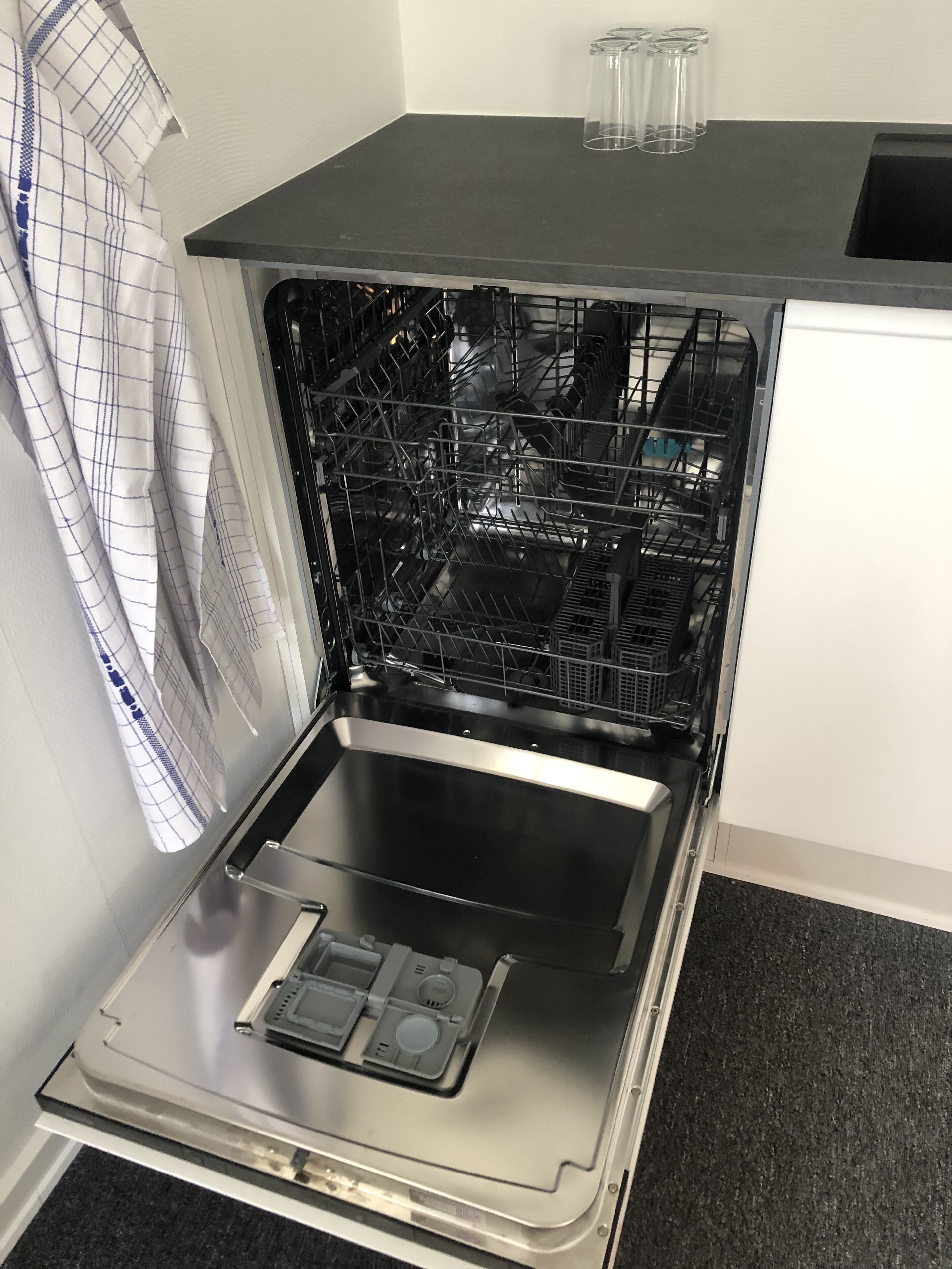 Un lave-vaisselle ouvert dans une cuisine avec un plan de travail noir et un mur blanc.