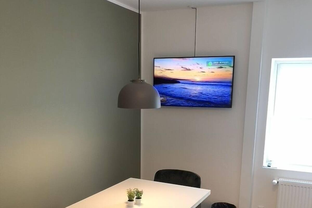 A small, modern room with a white table, two chairs, a TV mounted on the wall, and a window on the right.