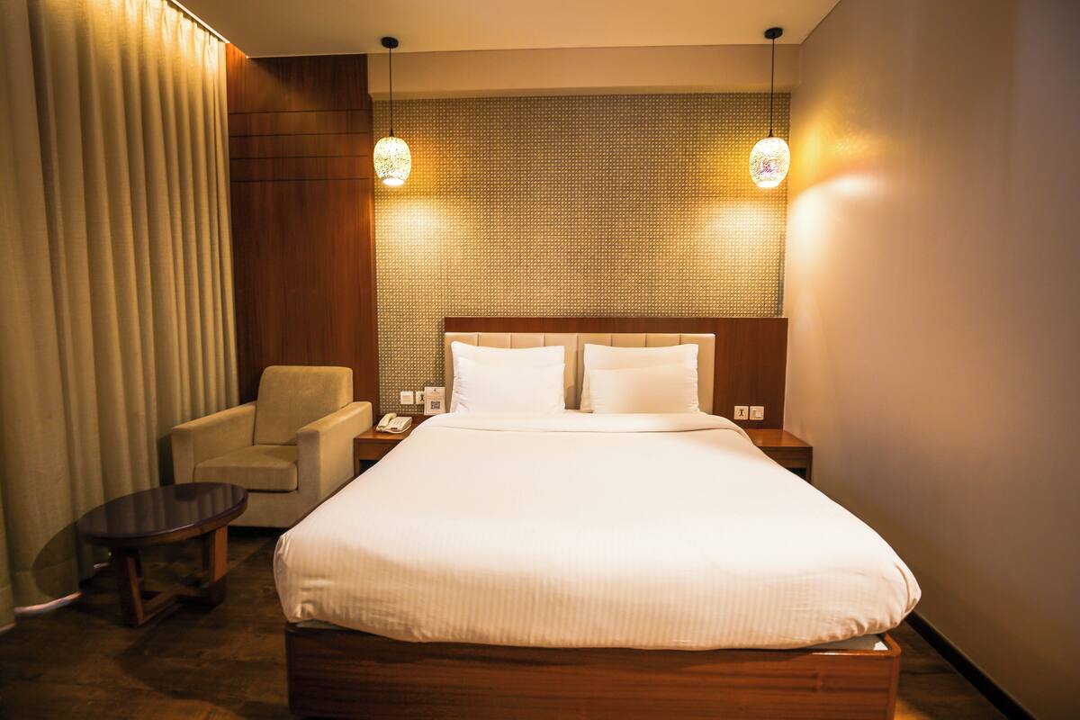 A hotel room with a large bed, a chair, a small table, and two wall-mounted lamps.
