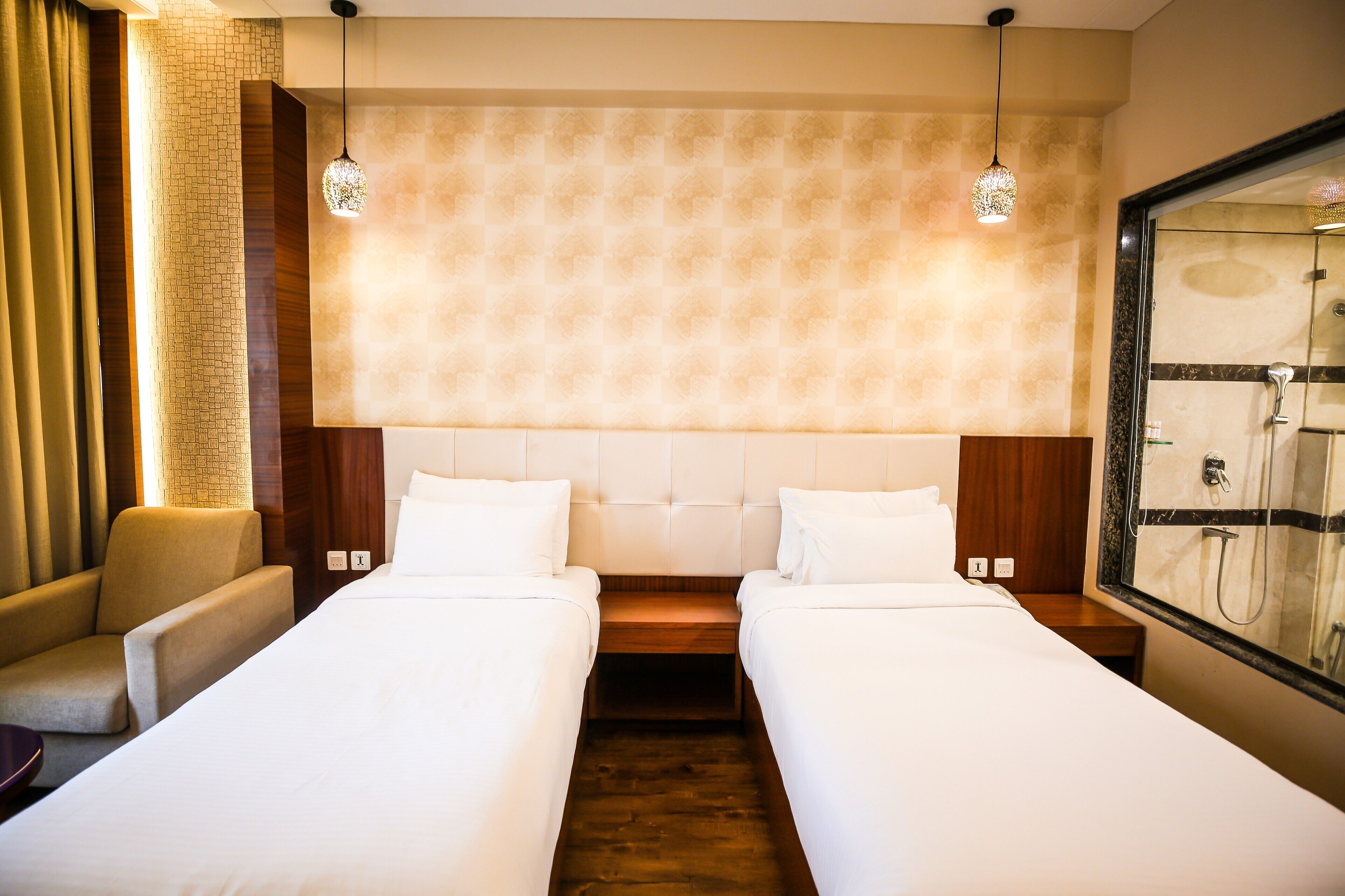 Habitación de hotel con dos camas individuales, un sofá, cabecero de madera y un espejo grande que refleja una zona de ducha.