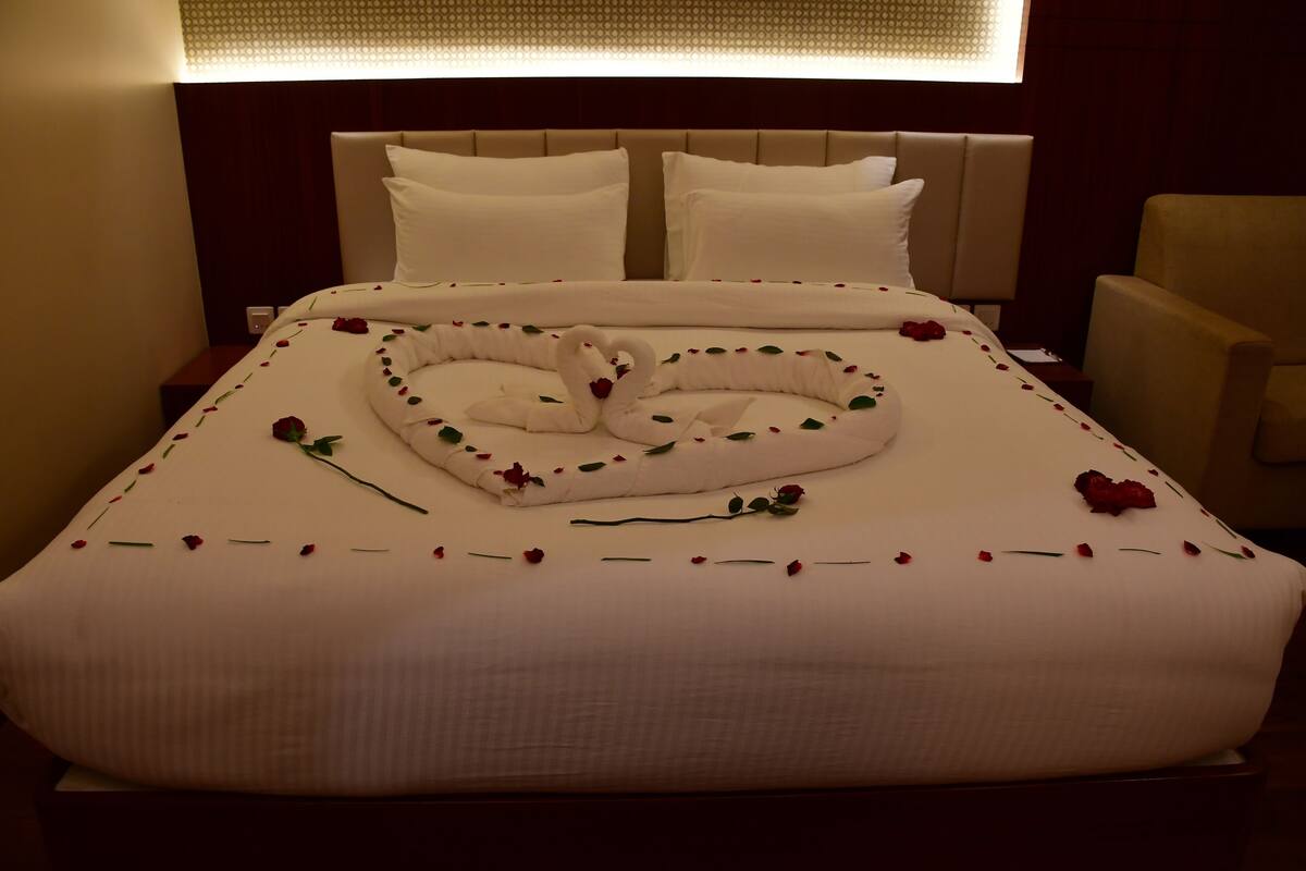 A neatly made bed with a heart-shaped towel and rose petals, a headboard, and a chair.