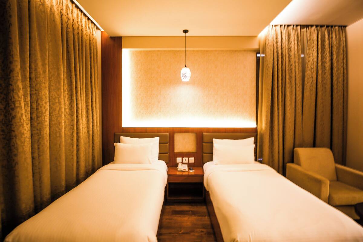 A hotel room with two single beds, a nightstand with a lamp, a chair, and a large window with curtains.