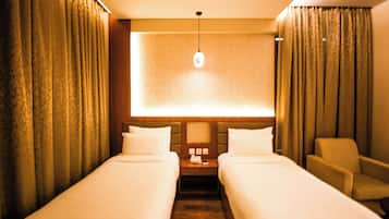 A hotel room with two single beds, a nightstand with a lamp, a chair, and a large window with curtains.