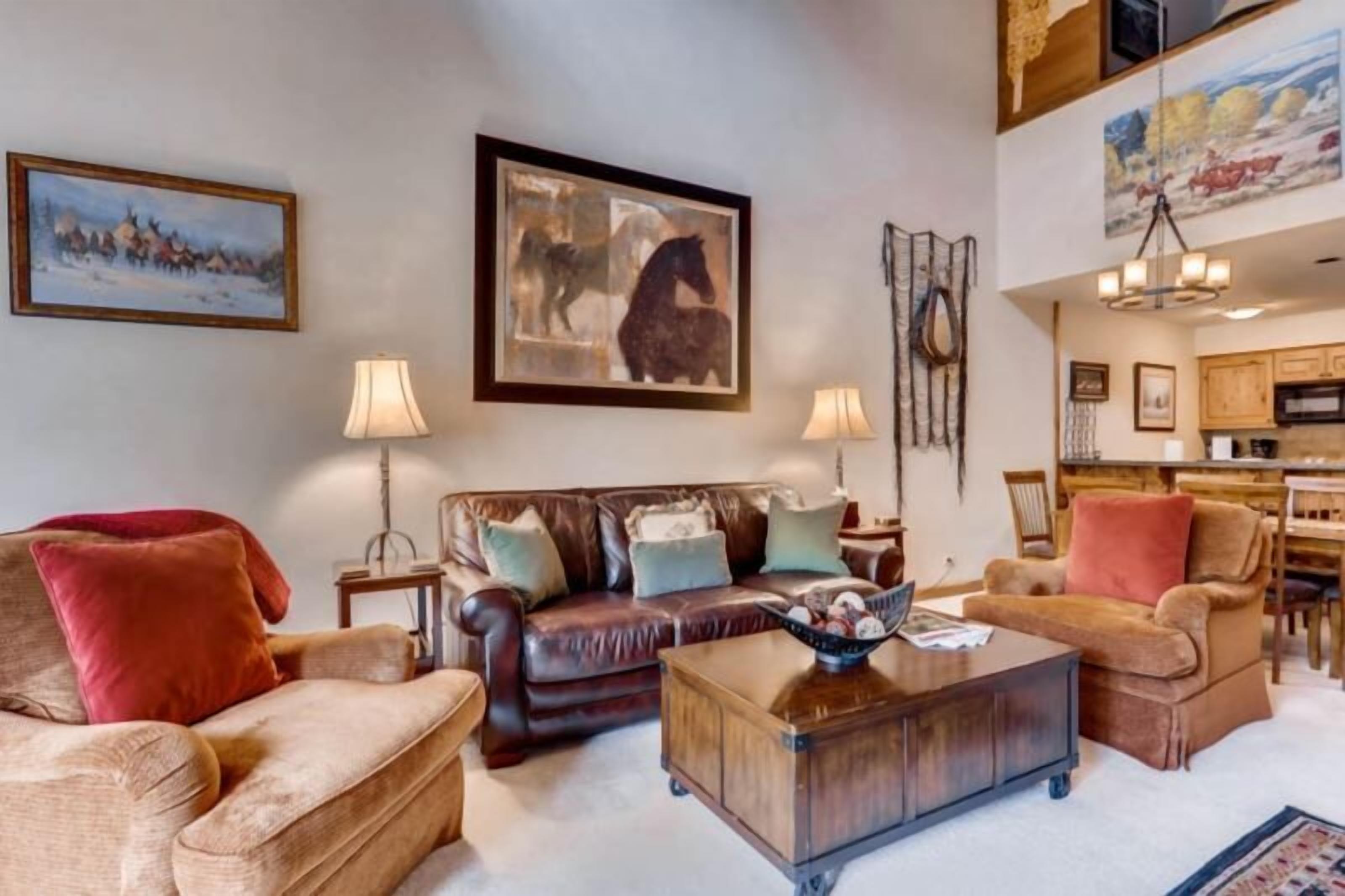 A living room with a brown leather sofa, a wooden coffee table, and two armchairs. There are framed paintings on the walls and a chandelier above the dining area.