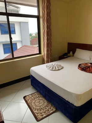 A hotel room with a bed, a window with a view of buildings, a patterned rug, and a decorative item on the bed.