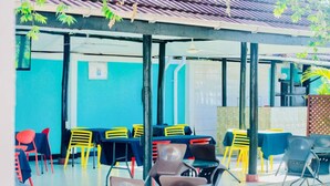 Un espace extérieur aménagé avec des tables et des chaises, une partie abritée et un bâtiment doté d’un toit en tuiles et de murs bleus.