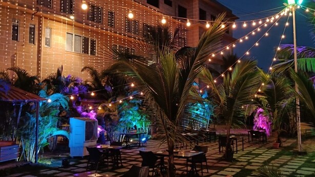 An outdoor patio with string lights, palm trees, and a building in the background. - TGN SUITS - HOTEL (Raipur)