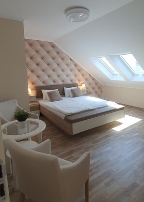 A modern bedroom with a tufted headboard, a bed with white bedding, a small round table with a plant, and a seating area with chairs. - Camille Apartments (Budapest)