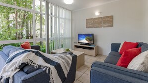 Una sala de estar con televisor, sofá, mesa de centro y vistas a zonas verdes a través de amplios ventanales.