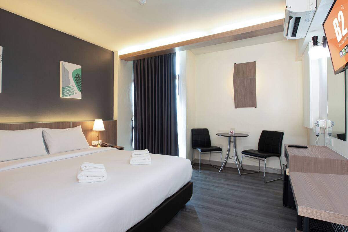 Ein Hotelzimmer mit einem großen Bett, zwei Nachttischlampen, einem kleinen Tisch mit Stühlen, einem Fön und einem Fernseher, der die Zimmernummer B2 anzeigt.