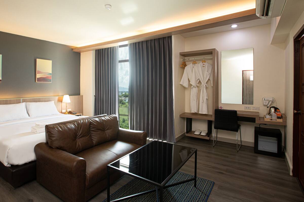 Ein modernes Hotelzimmer mit einem Kingsize-Bett, einem Ledersofa, einem Glastisch, einem Schreibtisch mit Stuhl, einem Bademantelständer und einem großen Fenster mit Vorhängen.