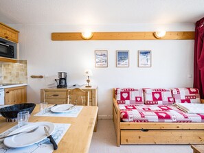 Een compacte kamer met een kitchenette, een eettafel met bestek, een bed met een decoratief deken en drie ingelijste foto's aan de muur.