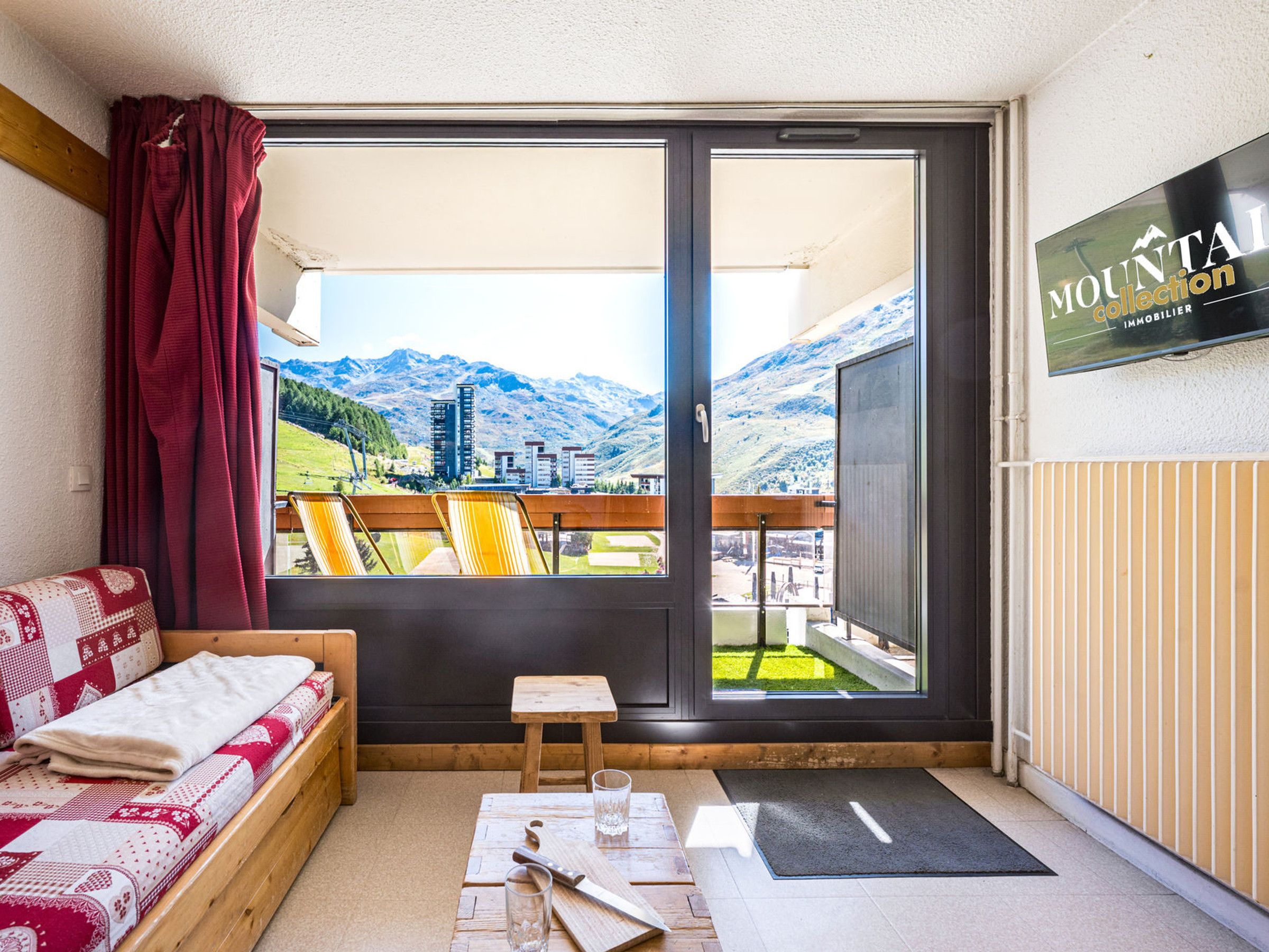 A room with a bed, a small table, a bench, and a view of mountains through a large sliding door.