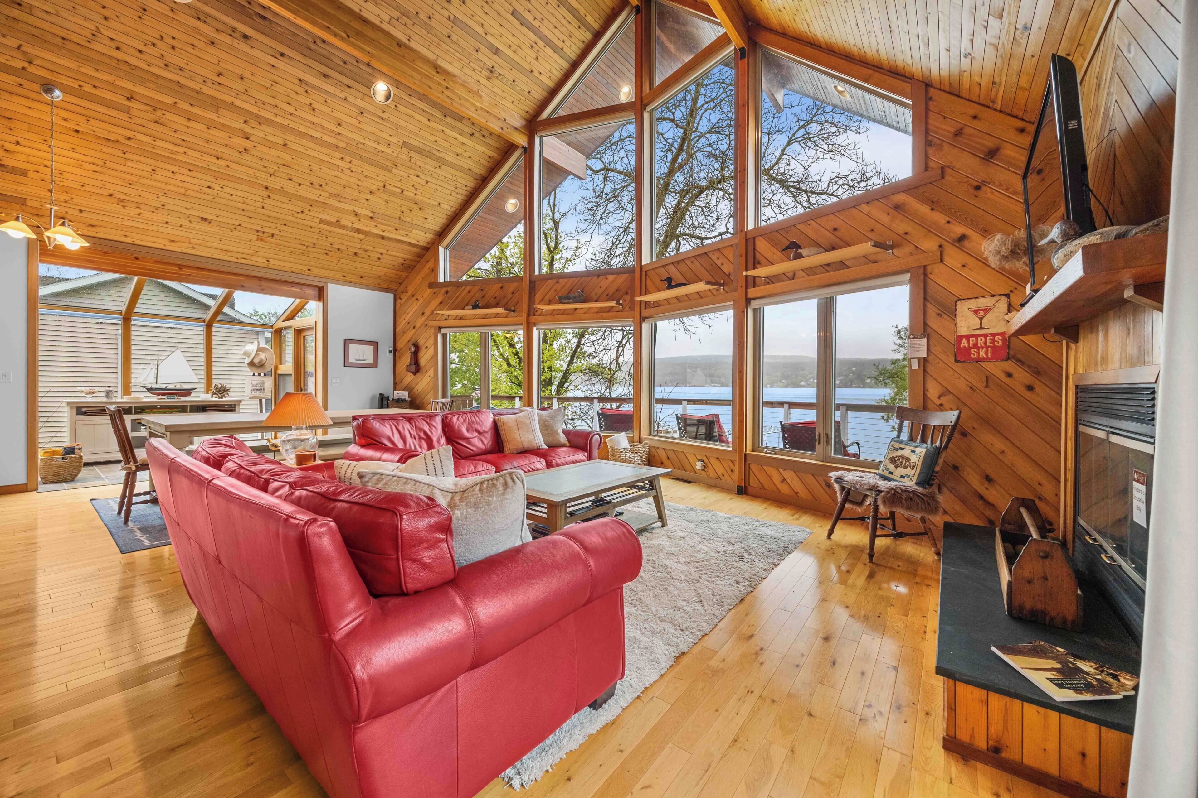 Une cabane spacieuse avec un grand plafond en bois, un canapé en cuir rouge, une cheminée et une vue sur un lac.