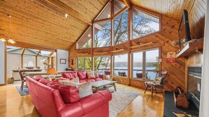 Une cabane spacieuse avec un grand plafond en bois, un canapé en cuir rouge, une cheminée et une vue sur un lac.