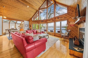 Une cabane spacieuse avec un grand plafond en bois, un canapé en cuir rouge, une cheminée et une vue sur un lac.