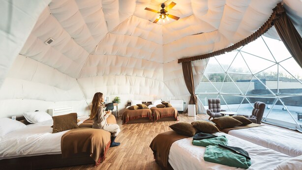 Ein gemütliches, gut beleuchtetes Zelt-ähnliches Zimmer mit mehreren Betten, einem Deckenventilator und großen Fenstern mit Blick ins Freie.
