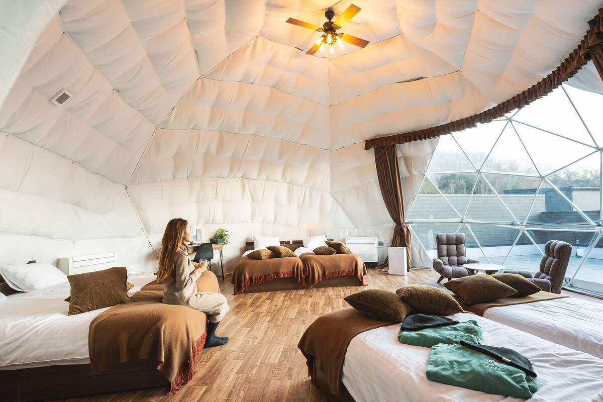 Ein gemütliches, gut beleuchtetes Zelt-ähnliches Zimmer mit mehreren Betten, einem Deckenventilator und großen Fenstern mit Blick ins Freie.