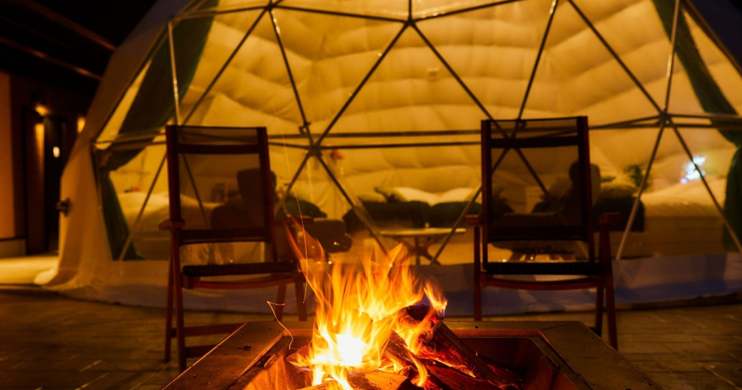 A geodesic dome structure with a fire pit in the center and two chairs facing it.