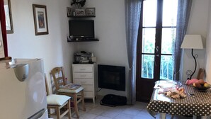 Een knus woongedeelte met een kitchenette, een bank, een eettafel met een tafelkleed met stippen, een televisie en een raam met gordijnen.