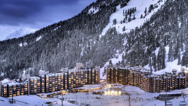En snötäckt semesterort med flera höga byggnader, skidliftar och en bergig bakgrund.
