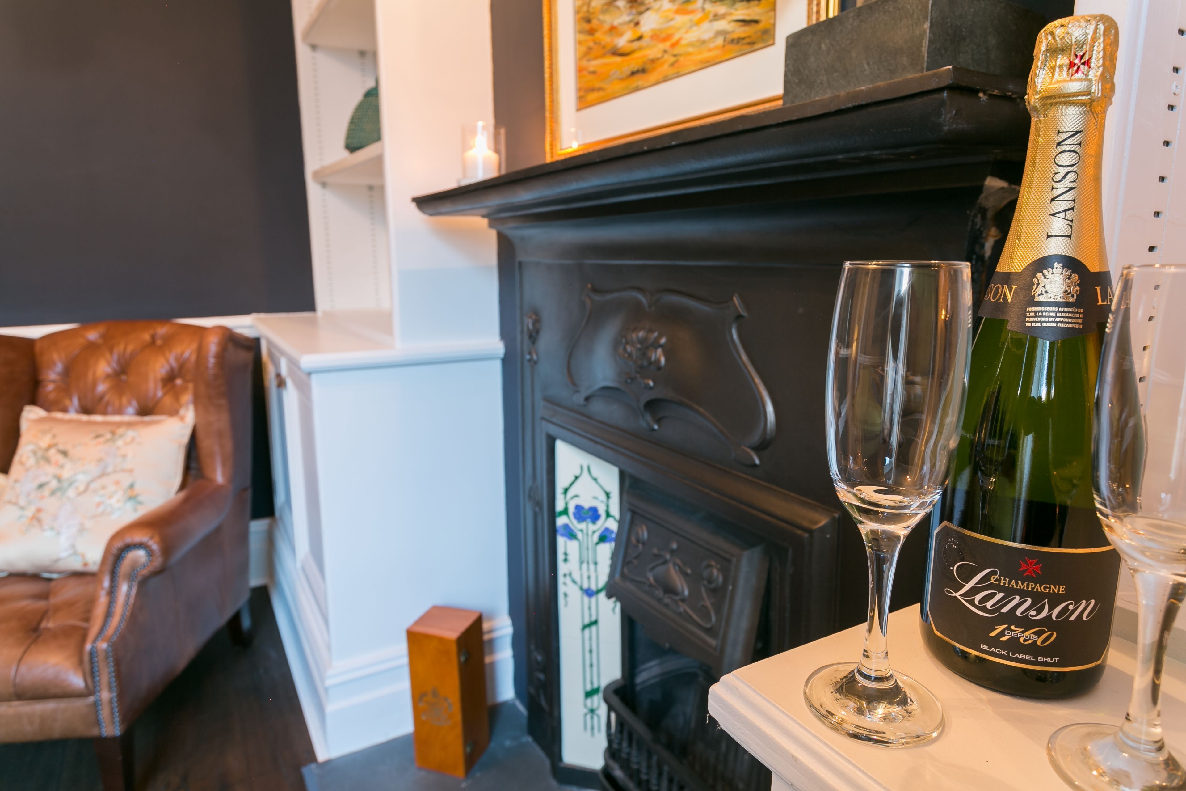 A bottle of champagne and two glasses on a table in a room with a fireplace, a leather armchair, and a decorative mirror.