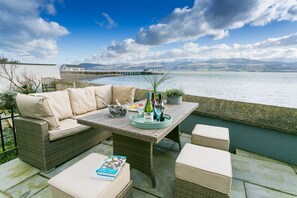 A patio with wicker furniture, a table set with a bottle, glasses, and a tray, overlooking a body of water and mountains. - Craig Hyfryd -  a coastal that sleeps 12 guests  in 6 bedrooms (Beaumaris)