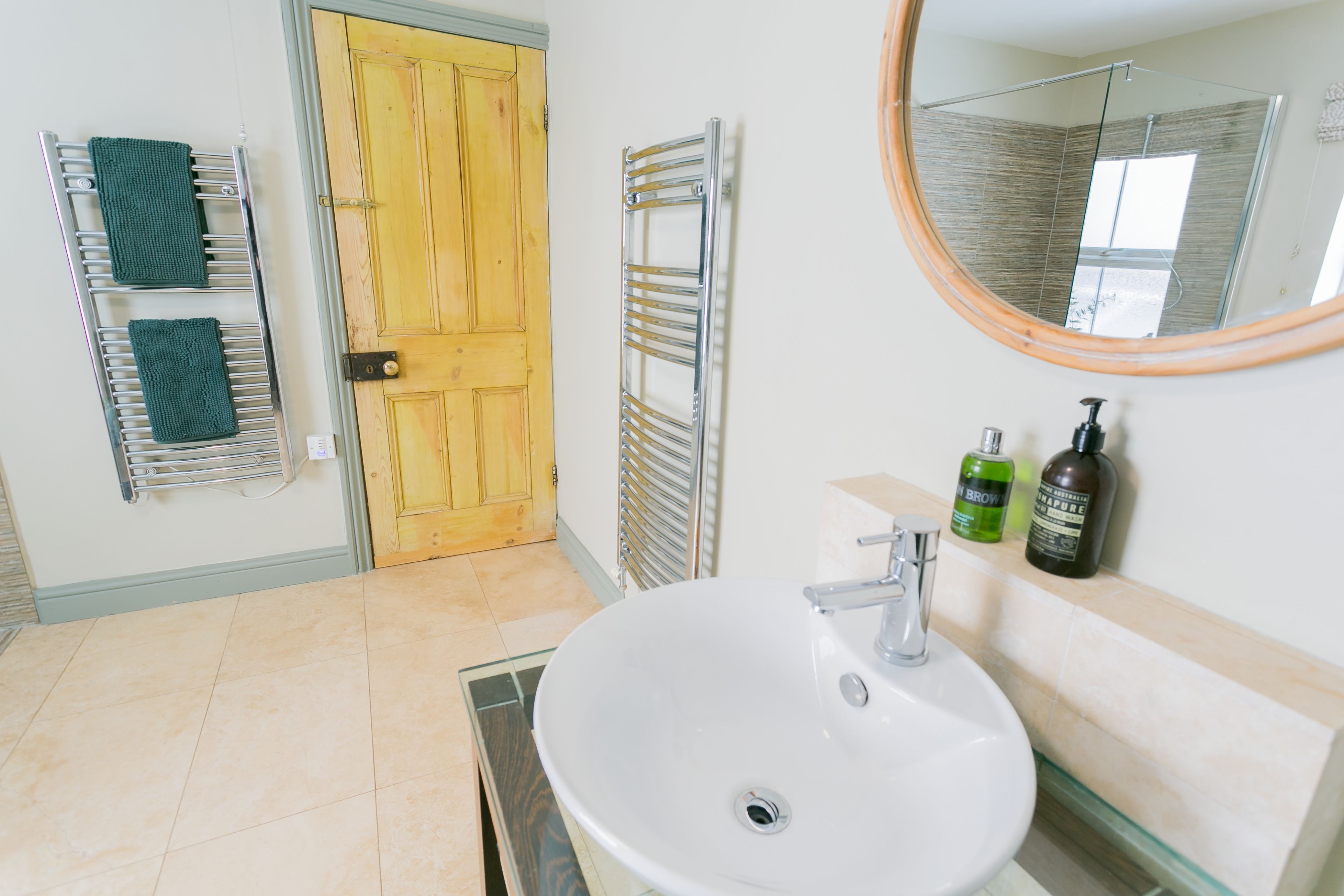 Un bagno con un lavandino bianco, una porta in legno, un porta asciugamani con asciugamani verdi, una cabina doccia in vetro e uno specchio.