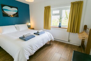 A bedroom with a bed, a window with curtains, a radiator, a wooden floor, and a wall with a heart-shaped artwork. - Cwter Hywel -  a cosy fire that sleeps 6 guests  in 3 bedrooms (Llandrygarn)