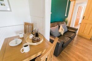 A dining area with a wooden table, plates, cups, and a cake. A brown leather sofa and a green wall are in the background. - Cwter Hywel -  a cosy fire that sleeps 6 guests  in 3 bedrooms (Llandrygarn)