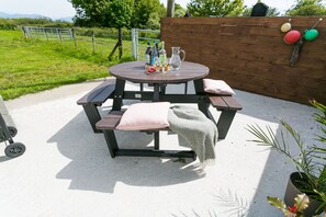 A round wooden table with benches, a picnic table, and a wooden fence in the background. - Cwter Hywel -  a cosy fire that sleeps 6 guests  in 3 bedrooms (Llandrygarn)